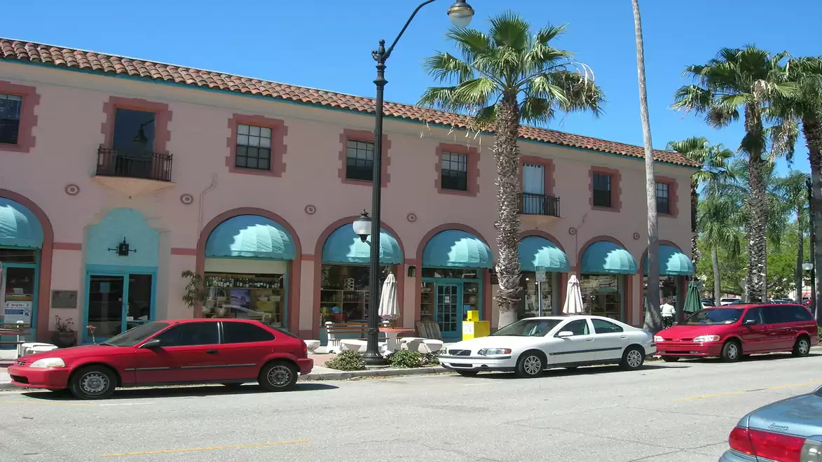 A pastel-colored building in Mediterranean style with blue awnings. Cars are parked along the palm-lined street.