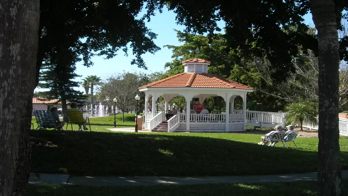 A picturesque gazebo with a red roof stands in the middle of a green park, surrounded by trees.