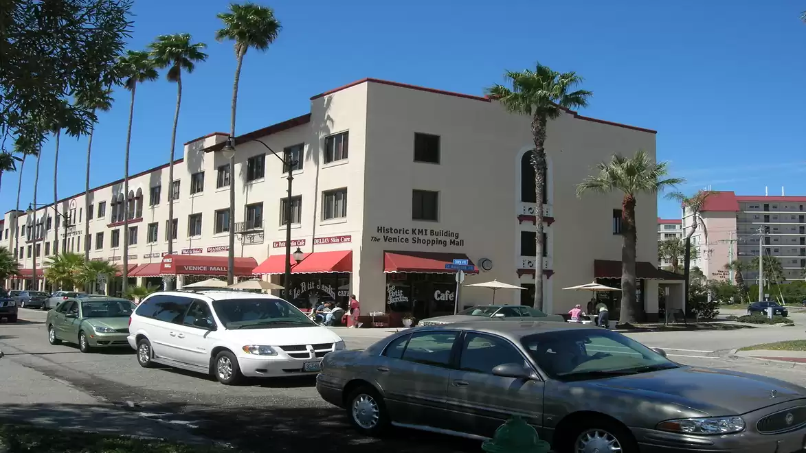 Street scene with modern vehicles in front of a shopping mall with palm trees and blue sky.