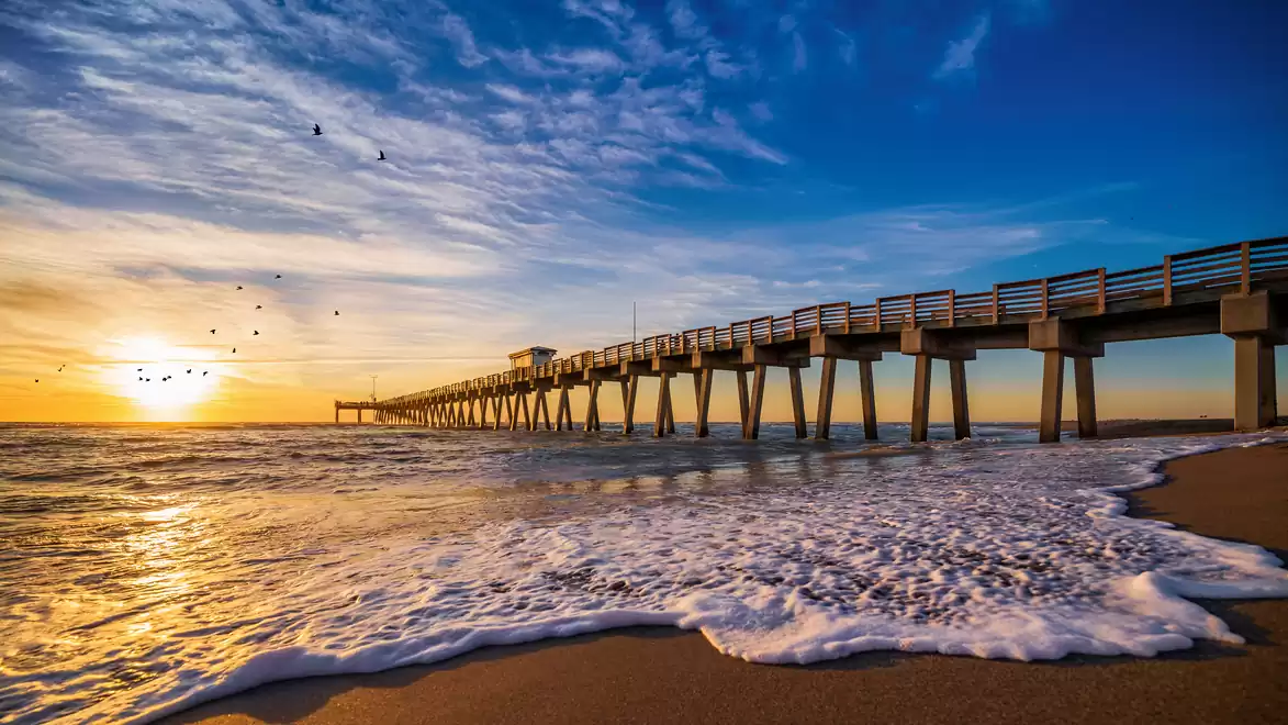 A long pier stretches over the sea under a dramatic sunset, the sky is filled with scattered clouds and flying birds.