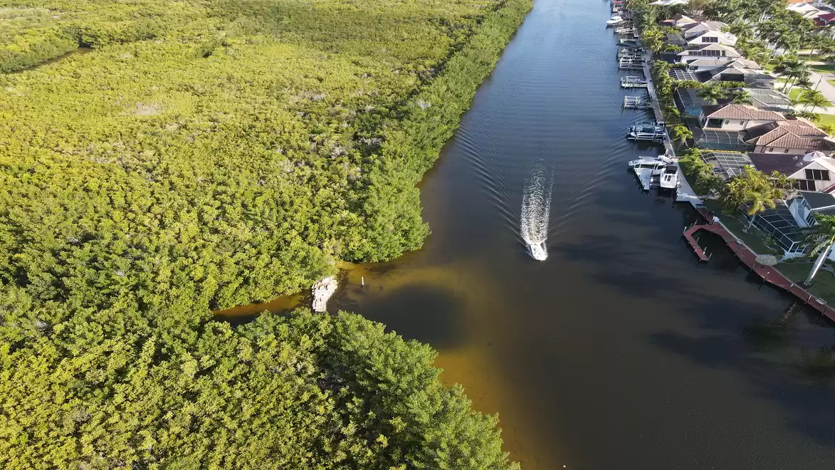 Aerial view of a canal with a boat navigating between green mangrove forest on the left and residential houses on the right.