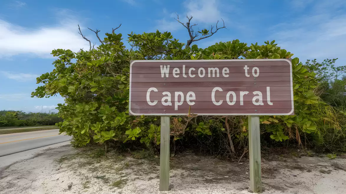 Welcome sign of Cape Coral in front of a green bush landscape under a blue sky.