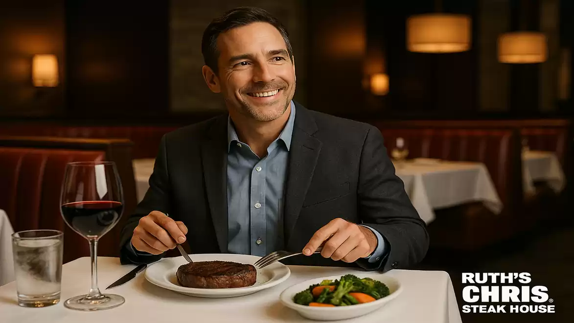 A smiling man in a suit sits at a restaurant table with a steak and vegetables on the plate.