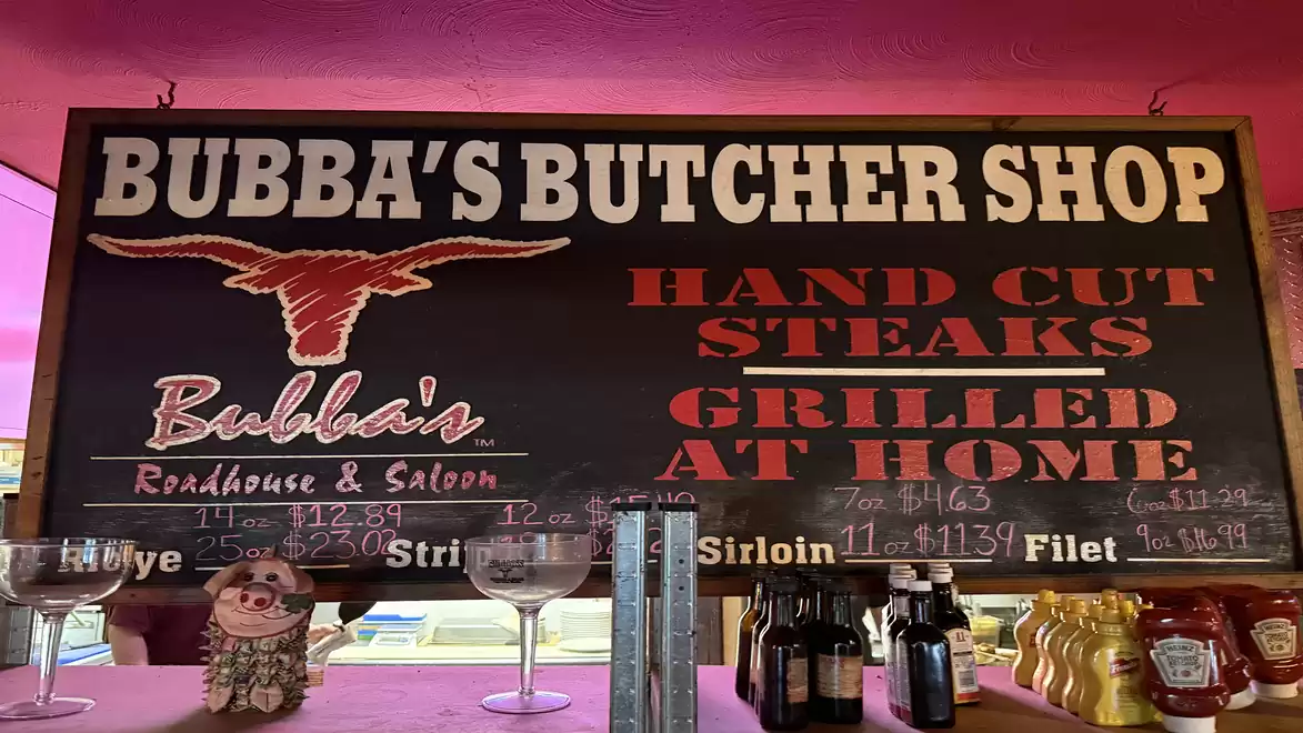 Sign for Bubba's Butcher Shop offering hand-cut steaks for grilling at home, decorated with glasses and sauces.