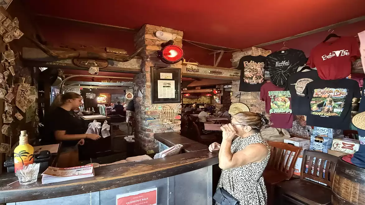 Cozy restaurant with wooden tables and decorations. A woman is standing at the counter and a waitress is visible in the background.