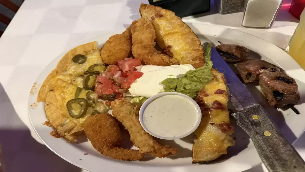 A plate of assorted finger foods, including nachos with cheese, jalapeños, guacamole, sour cream, and fried snacks. A large knife lies next to the plate.