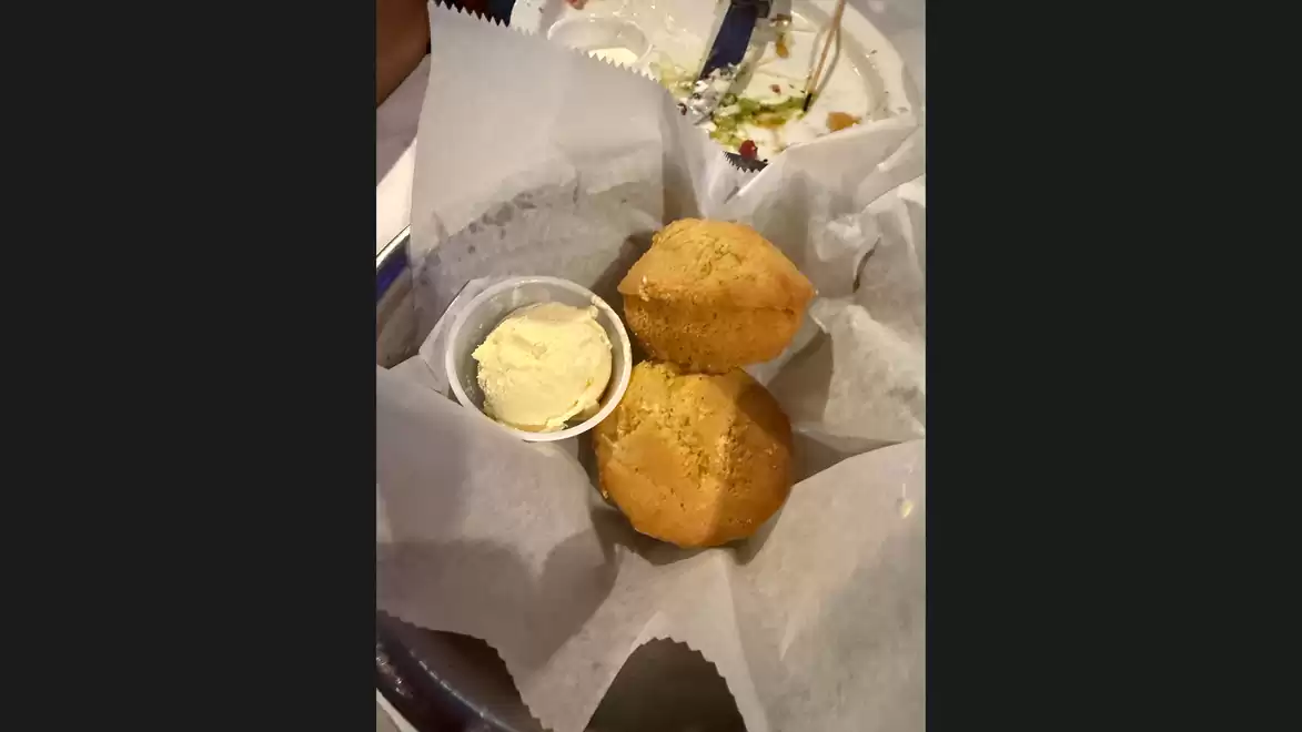 Two golden muffins in a paper basket, served with a portion of butter in a small cup.
