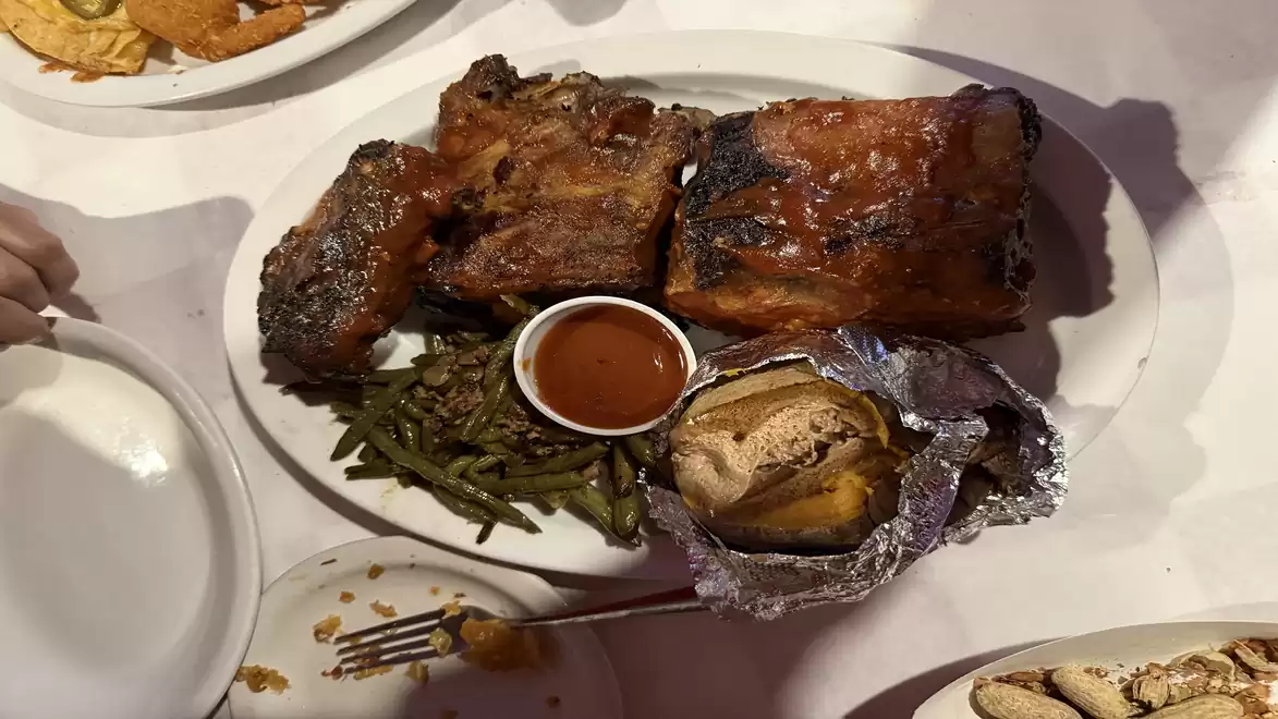 A plate with grilled ribs, green beans, a foil-wrapped potato, and a sauce dish.