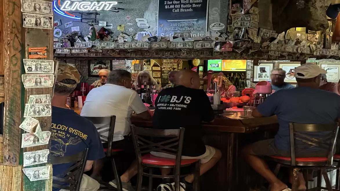 A group of people sitting at a bar decorated with dollar bills. TVs and neon lights are visible in the background.