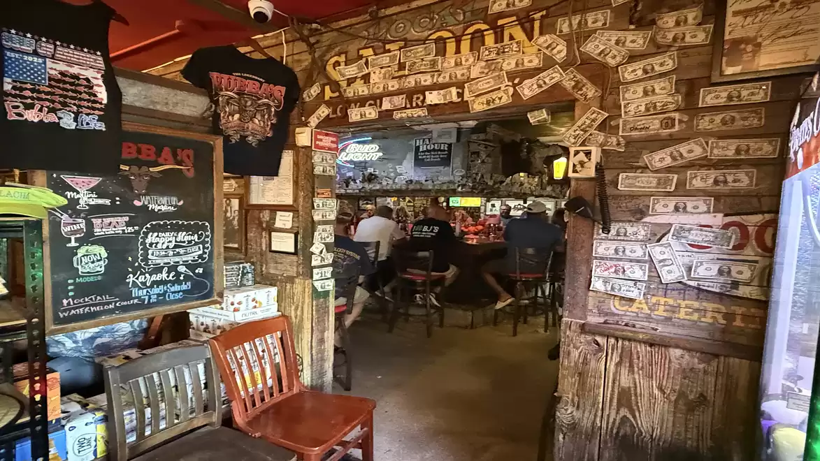 A rustic bar interior with dollar-adorned walls and wooden furniture. People are seated at the bar in the background.