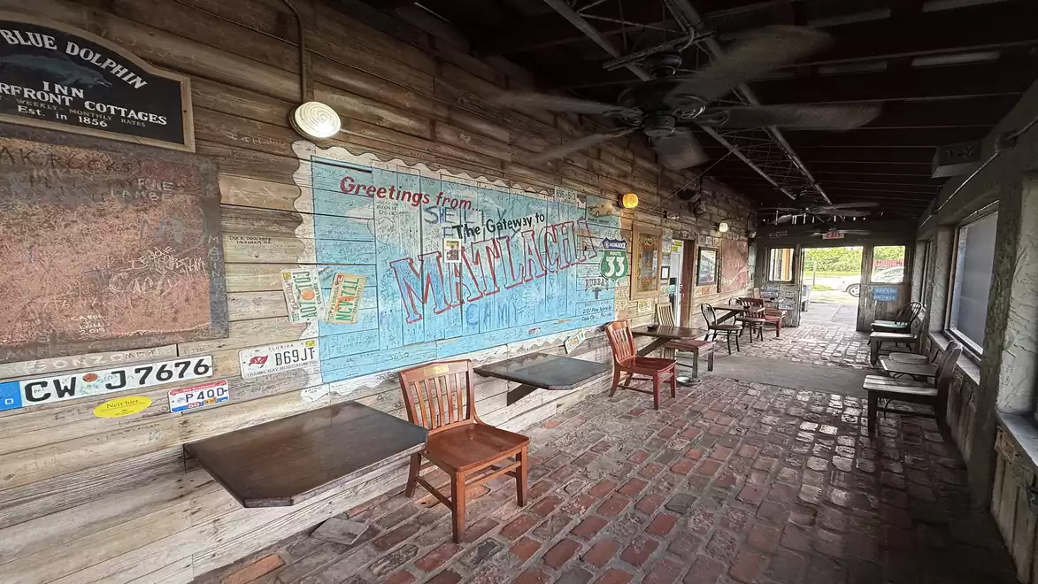 A rustic restaurant courtyard with wooden tables and chairs, brick flooring, and a wall mural reading 'Greetings from'.