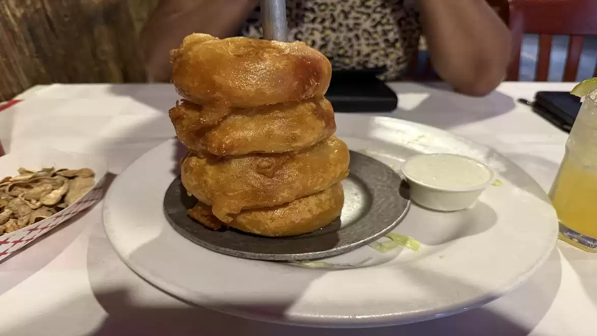 A plate with a tall stack of crispy onion rings next to a small bowl of dip and a drink in the background on a set table.