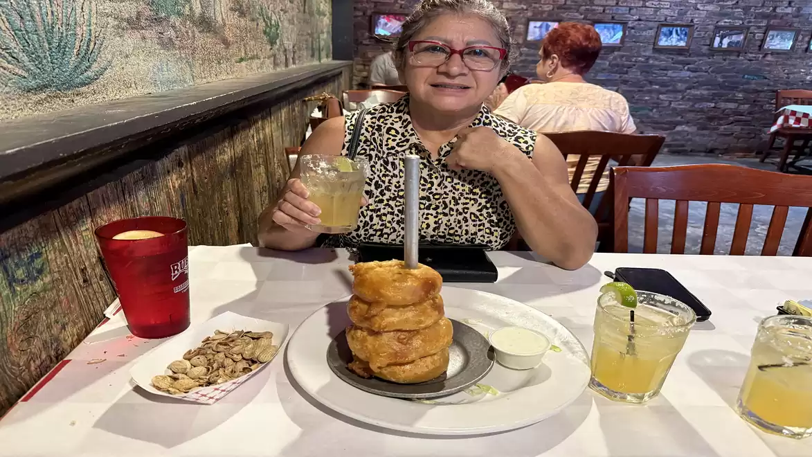 Older woman sitting in a restaurant enjoying a drink with a serving of onion rings and peanuts.