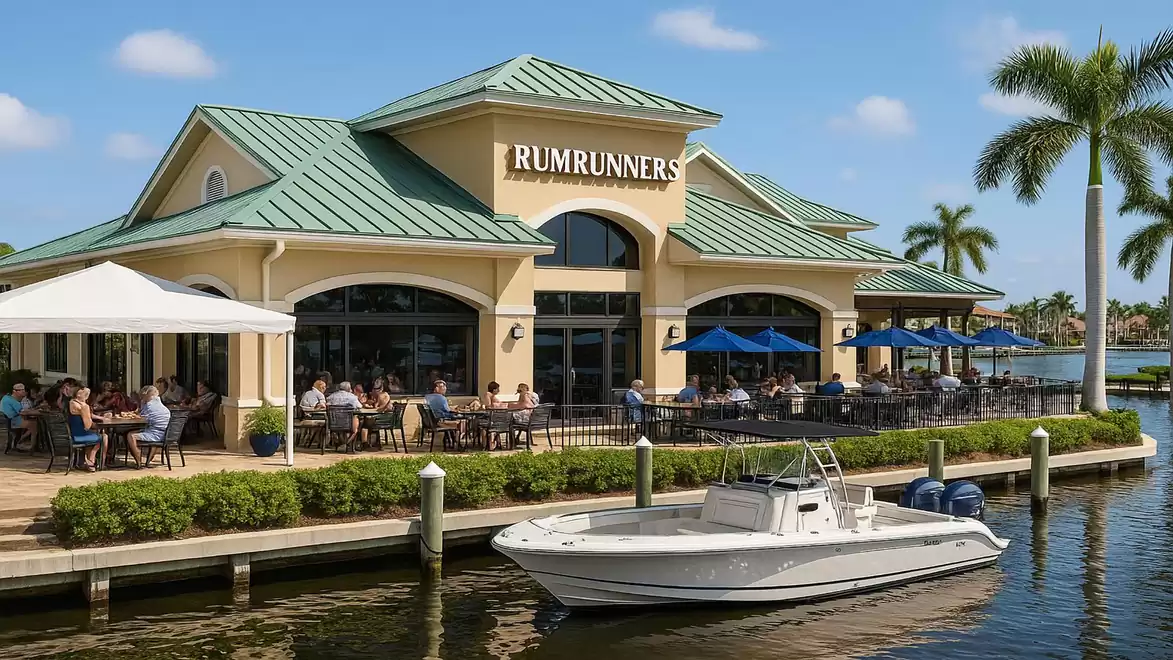 Waterfront restaurant with a boat docked nearby, surrounded by palm trees, sunny sky, and guests on the terrace.