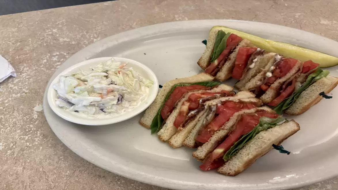 A plate with a multi-layered sandwich filled with tomatoes, lettuce, and meat, next to a bowl of coleslaw.