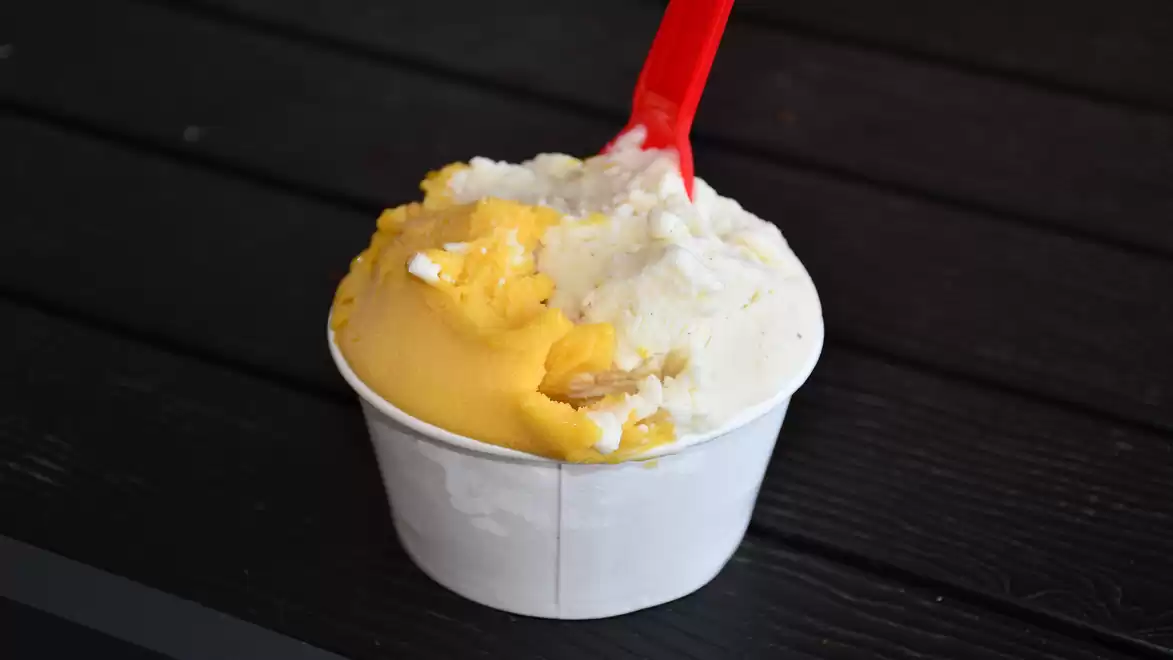 A cup of gelato ice cream in mango and vanilla flavors on a black-painted table, with a red plastic spoon.