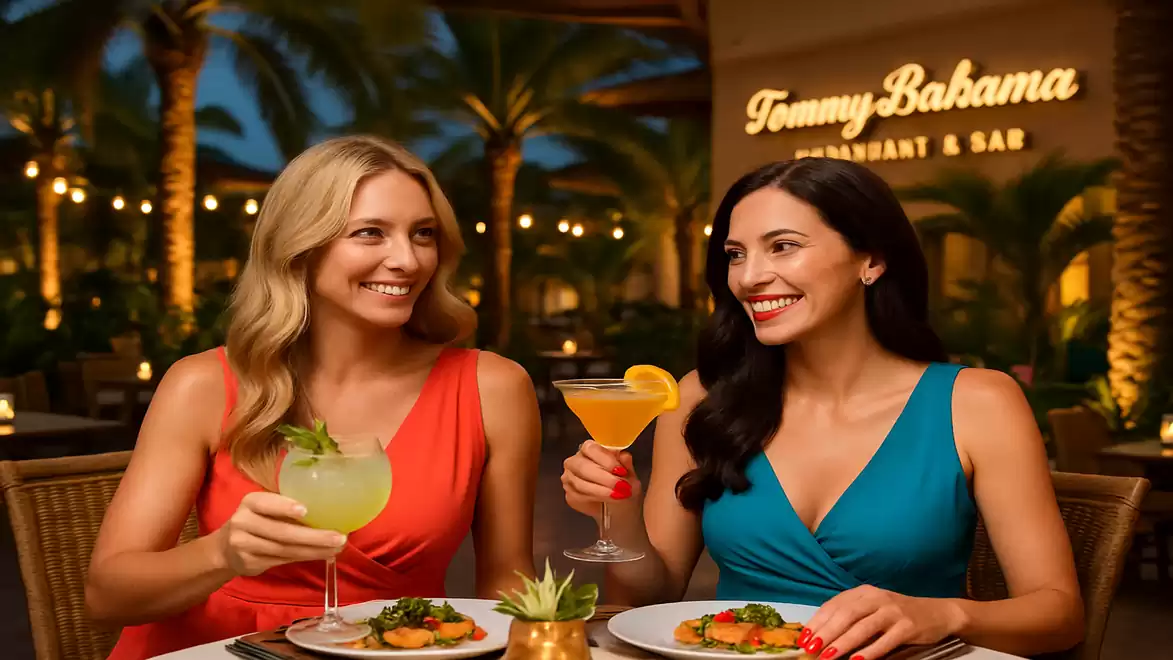 Two women in summer attire enjoying cocktails at a stylish restaurant with palm trees in the background.