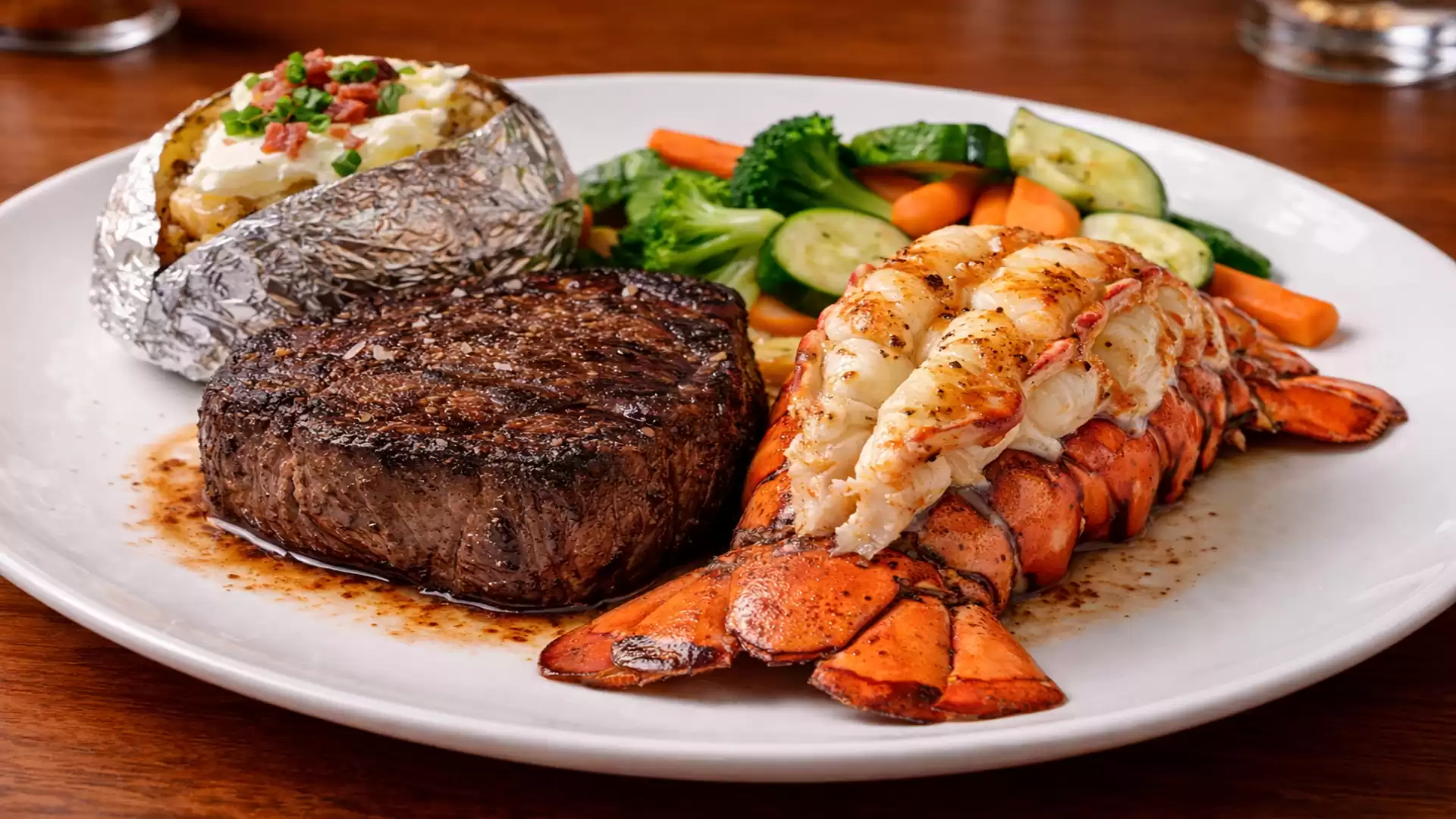 A plate with a juicy steak, a baked potato with sour cream and bacon, a grilled lobster tail half, and mixed vegetables including carrots, broccoli, and cucumbers.