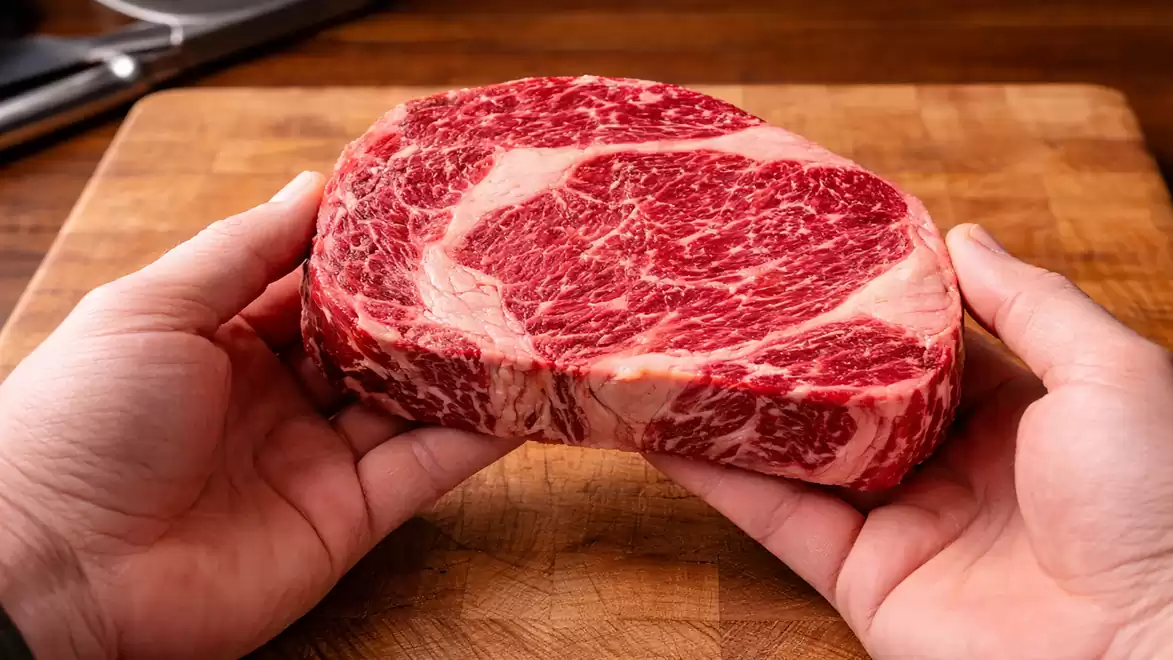 A person holds a raw, heavily marbled steak in both hands over a wooden board.