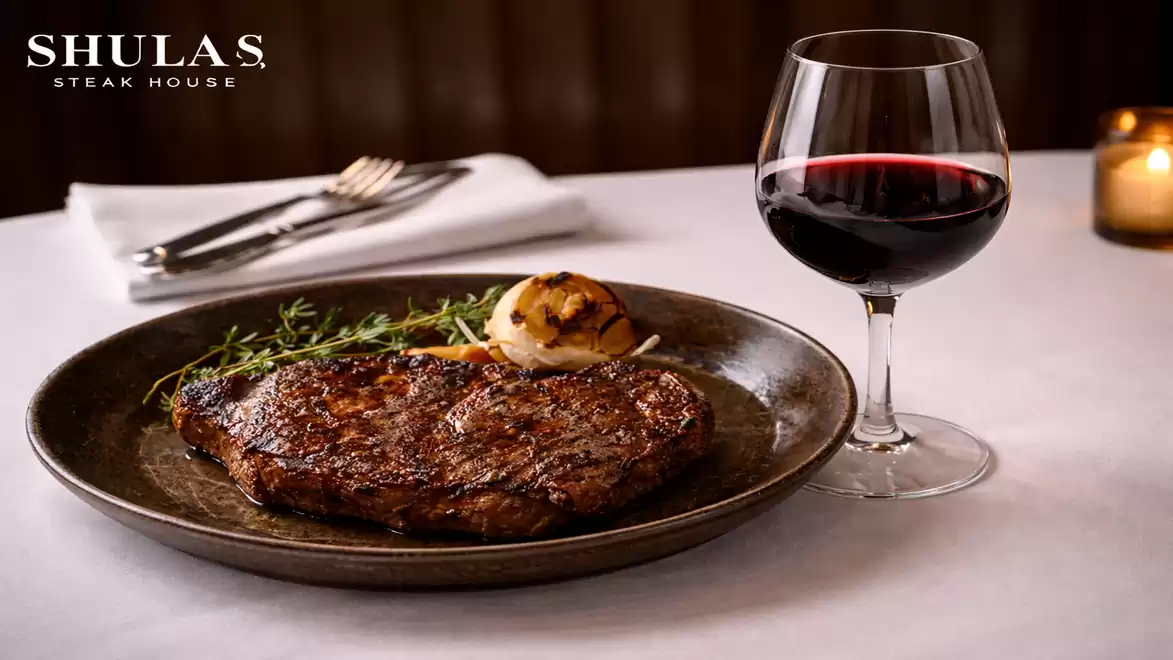 A juicy steak with grilled onions and fresh herbs, served on a white plate next to a glass of red wine. In the background, candlelight and cutlery are visible.