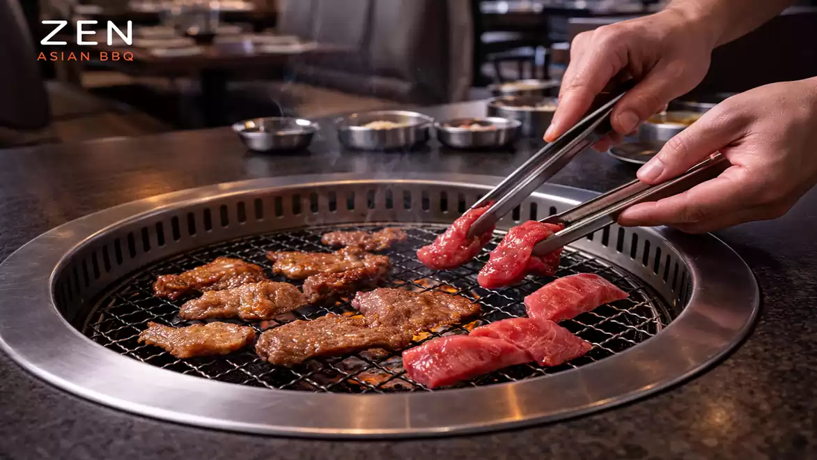 Close-up of a Korean BBQ grill with various meats being turned with tongs. Spice bowls are visible in the background.