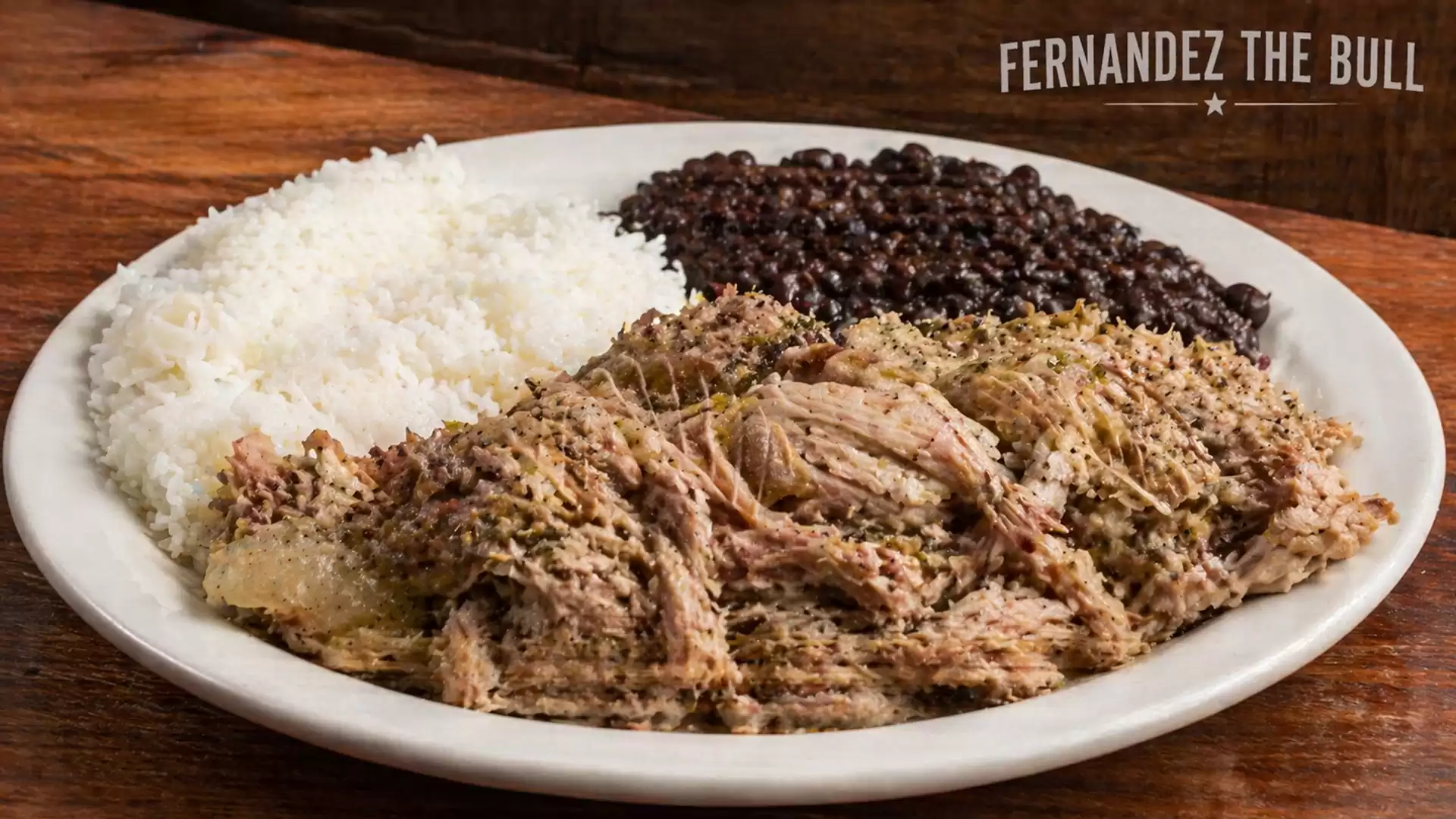 A plate with white rice, black beans, and tender, seasoned meat in front of a wooden background. 'Fernandez The Bull' is written in the background.