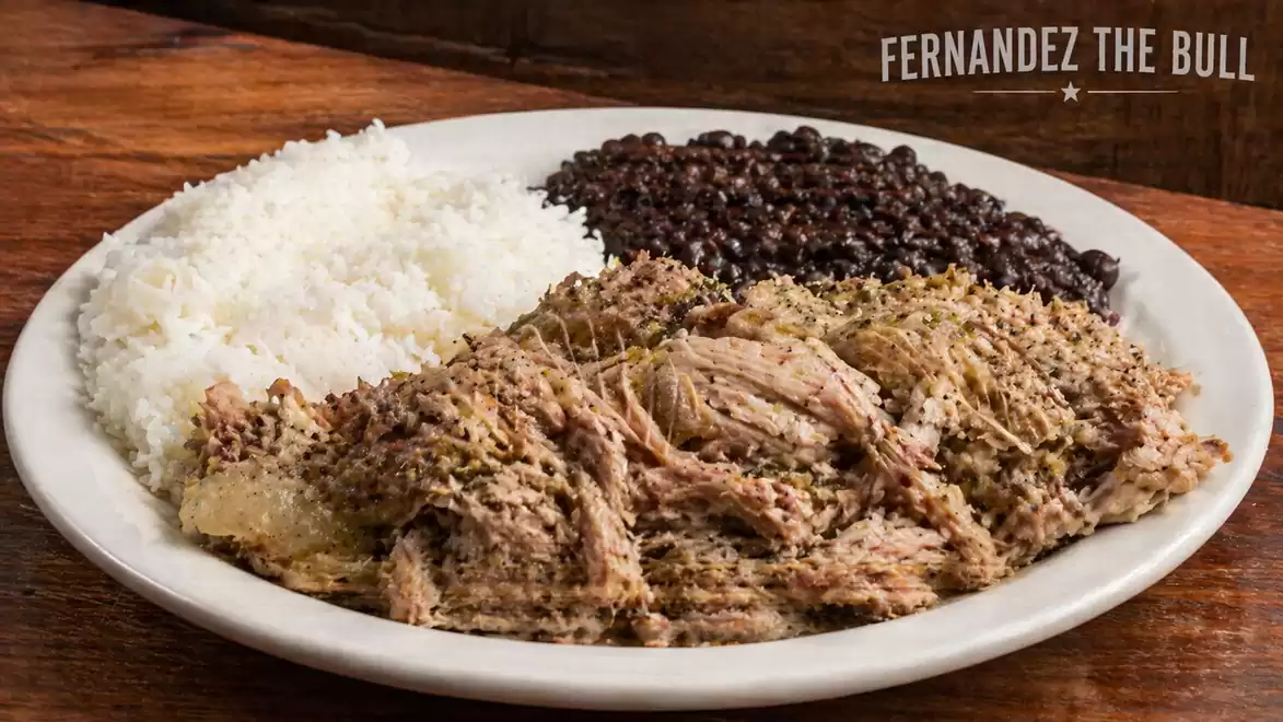 A plate with white rice, black beans, and tender, seasoned meat in front of a wooden background. 'Fernandez The Bull' is written in the background.