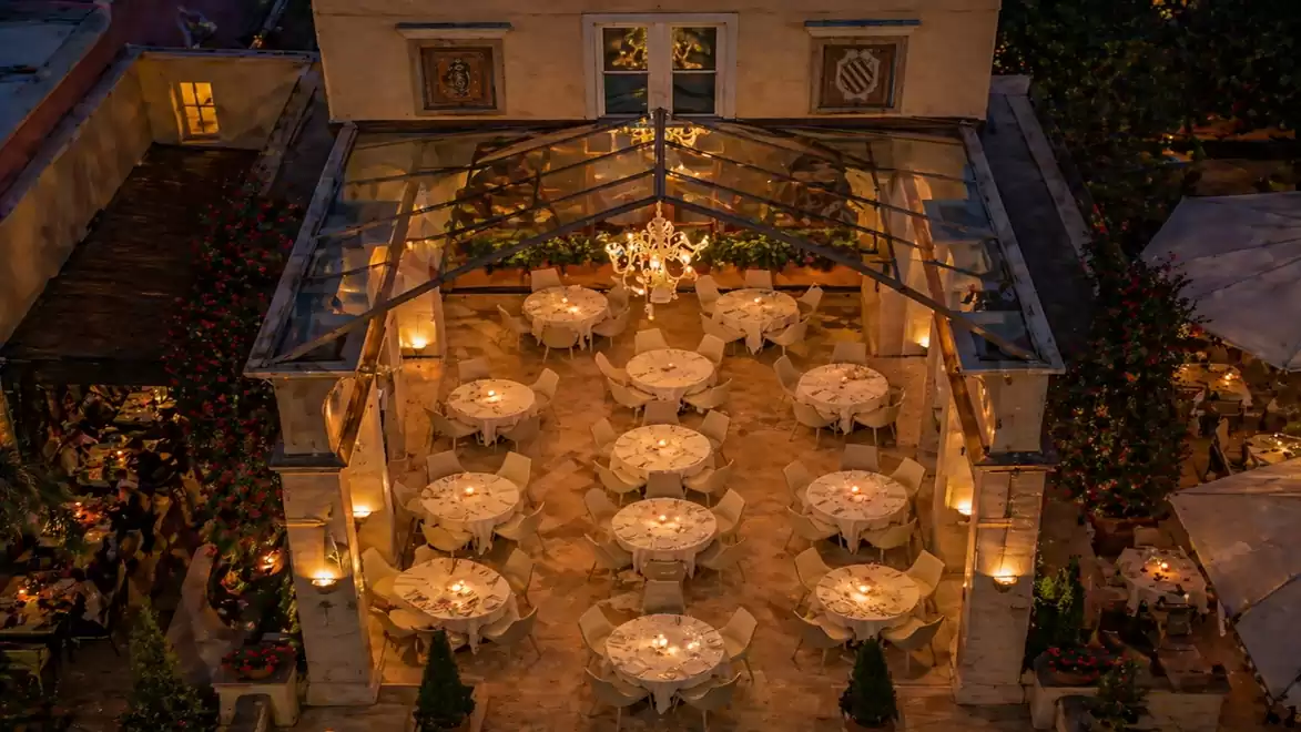 Aerial view of an elegantly lit restaurant with several round tables set for dinner under a glass pavilion.