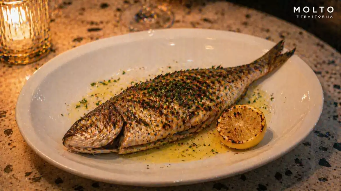 Grilled fish on oval plate with herbs and lemon slice on marbled table in a warmly lit restaurant.