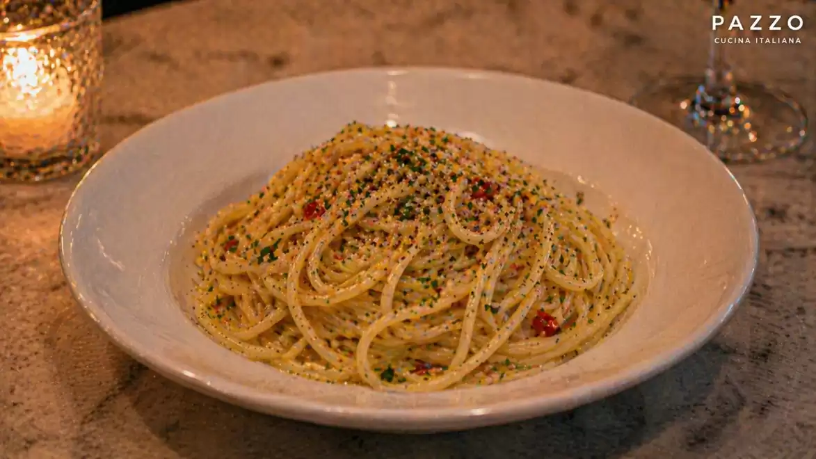 A plate of spaghetti served with cherry tomatoes and herbs on a dark table. A candle lights the background and a wine glass is beside it.