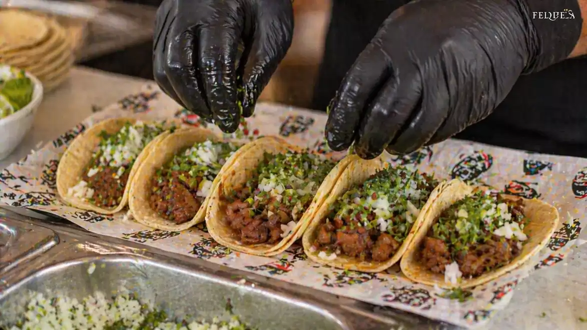 Close-up of hands in black gloves sprinkling green herbs on a row of tacos with meat and onions. The tacos are arranged on paper with a colorful pattern.