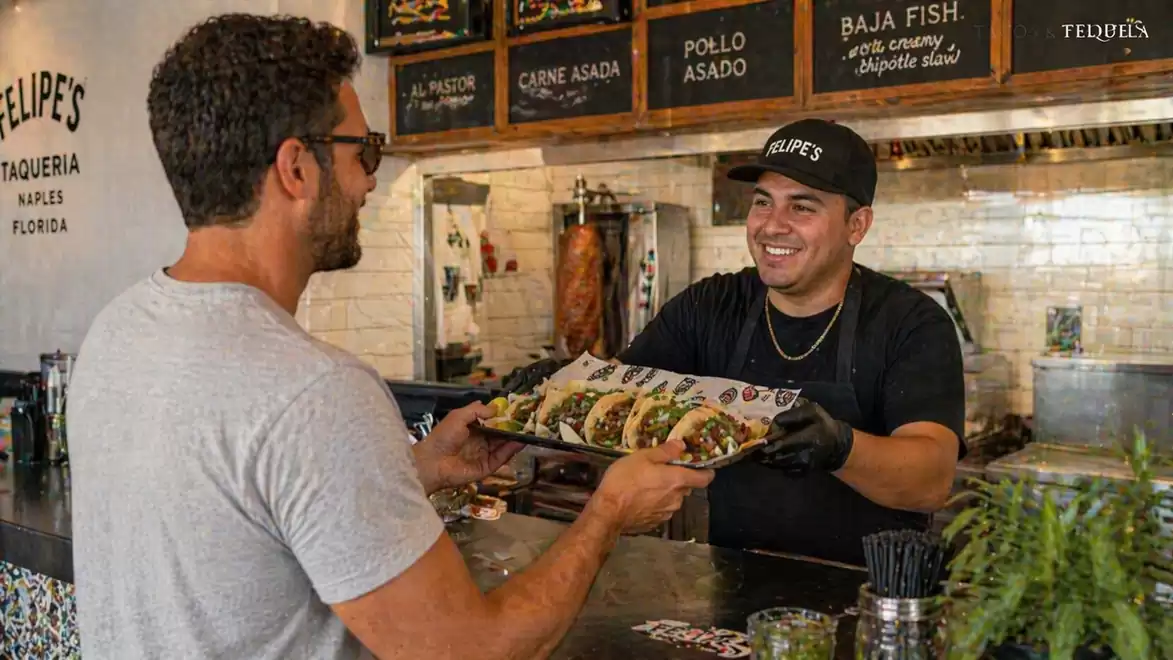 A man at a counter receives a tray of tacos from a smiling employee in a taqueria.