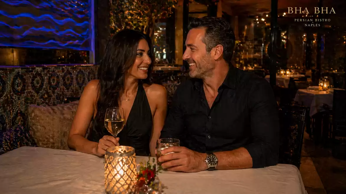 A couple sits at an elegantly set table in a moodily lit restaurant, smiling at each other. The table is decorated with a candle and a glass of wine. In the background, the warm ambiance of the BHA BHA Persian Bistro is visible.