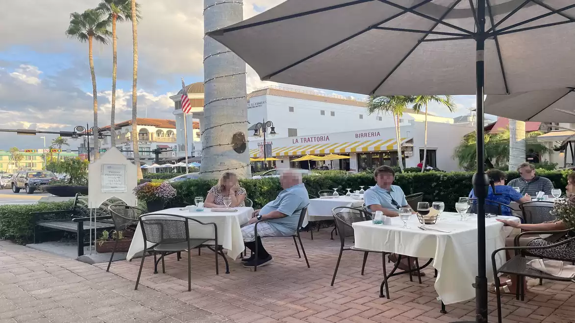 People sitting in a cozy street café with white tables, shaded by large umbrellas, surrounded by palm trees and a historic building in the background.