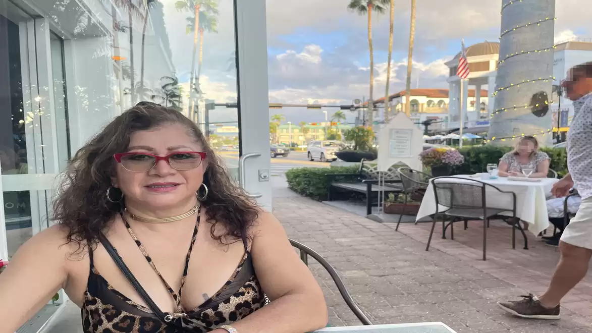 A woman with red glasses sits outside at a café, with a modern urban background.