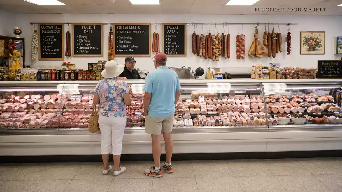 A man and a woman stand in front of a meat counter in a European food market. The counter is filled with various meats and sausages. Signs with product descriptions are hung on the wall.