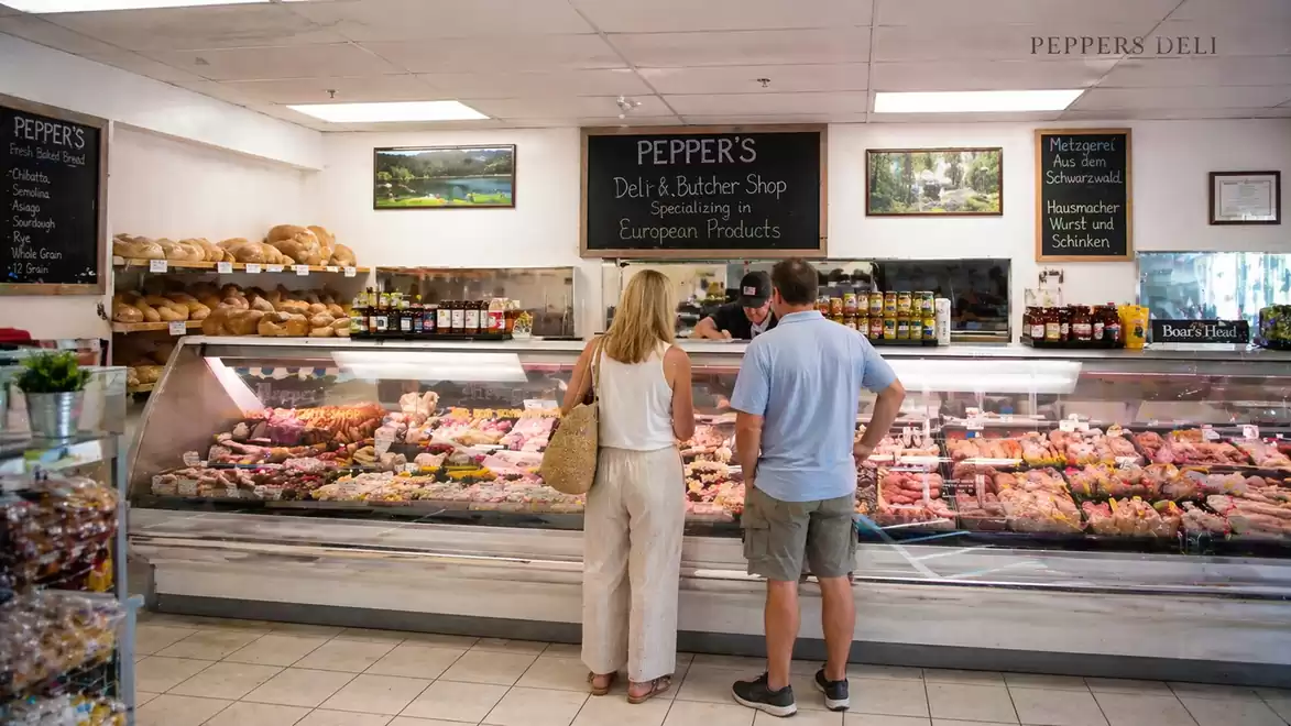 A woman and a man stand in a deli in front of a counter displaying a variety of meats and sausages. Shelves with bread and other products are visible in the background.