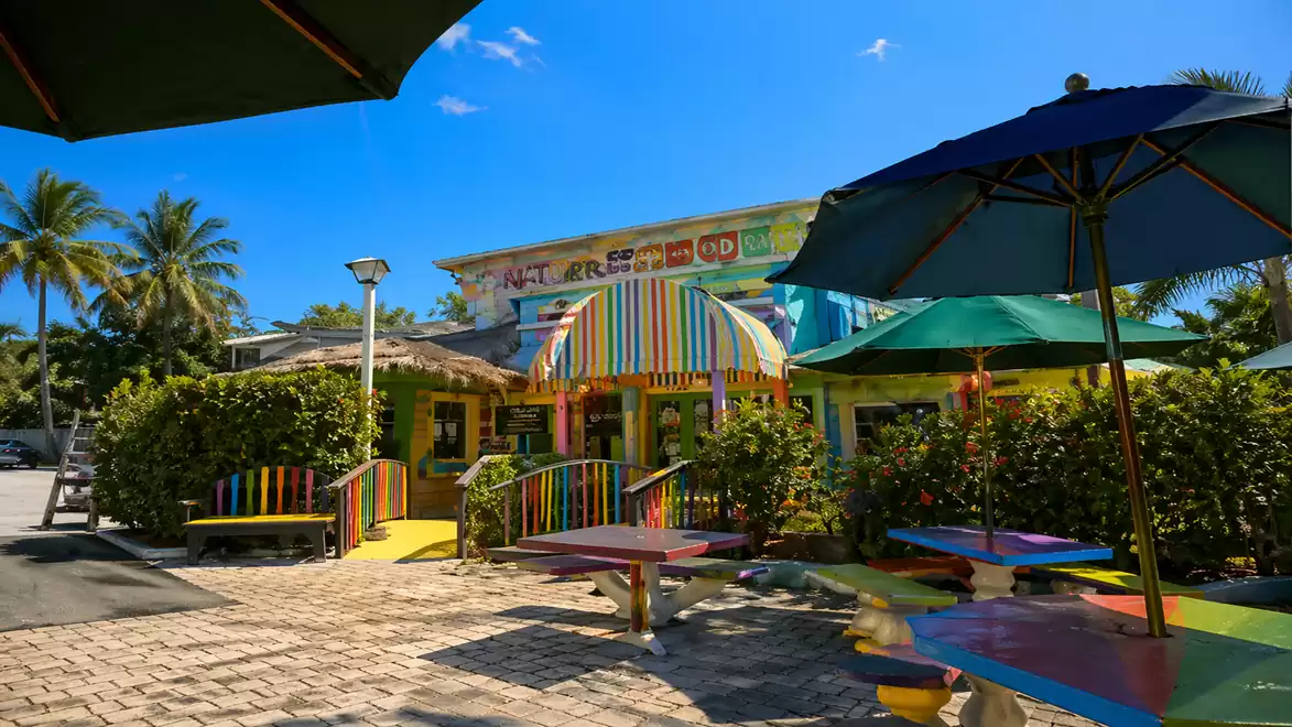 A colorful beach café with rainbow-colored awnings and tables, surrounded by palm trees in sunny weather.