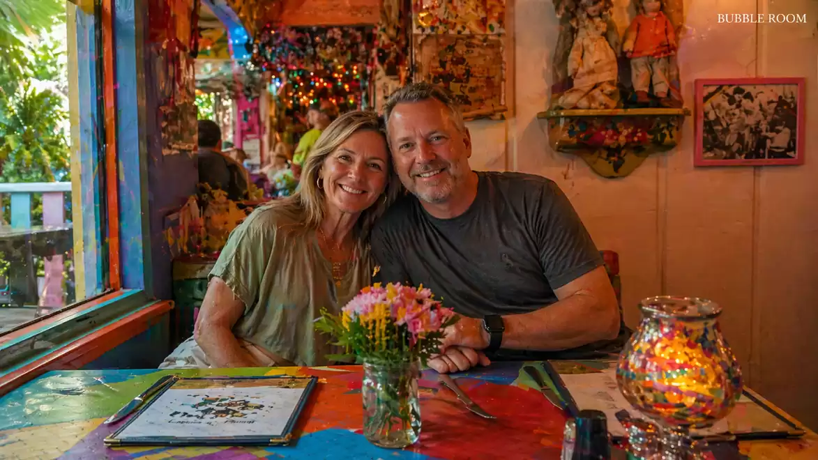 A smiling couple sits at a colorful table in a cozily decorated restaurant with a glowing lantern and flower arrangement.