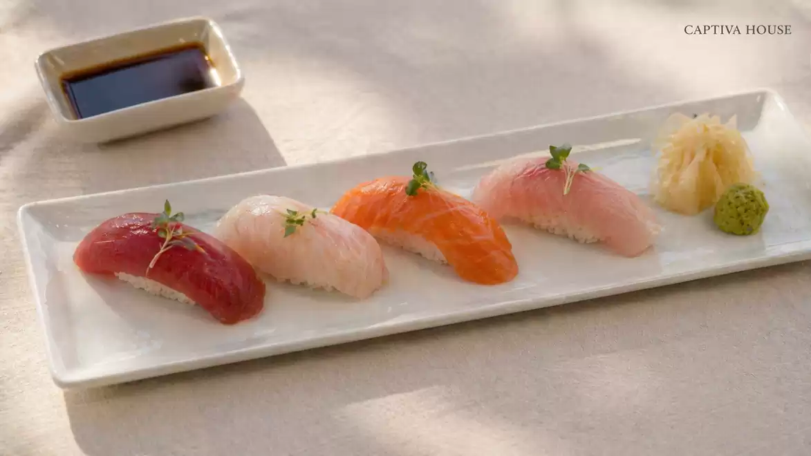 Sushi on a rectangular white plate with four different types of nigiri, garnished with small herbs. A dish with soy sauce is placed in the top left corner. Ginger and wasabi are visible in the background.