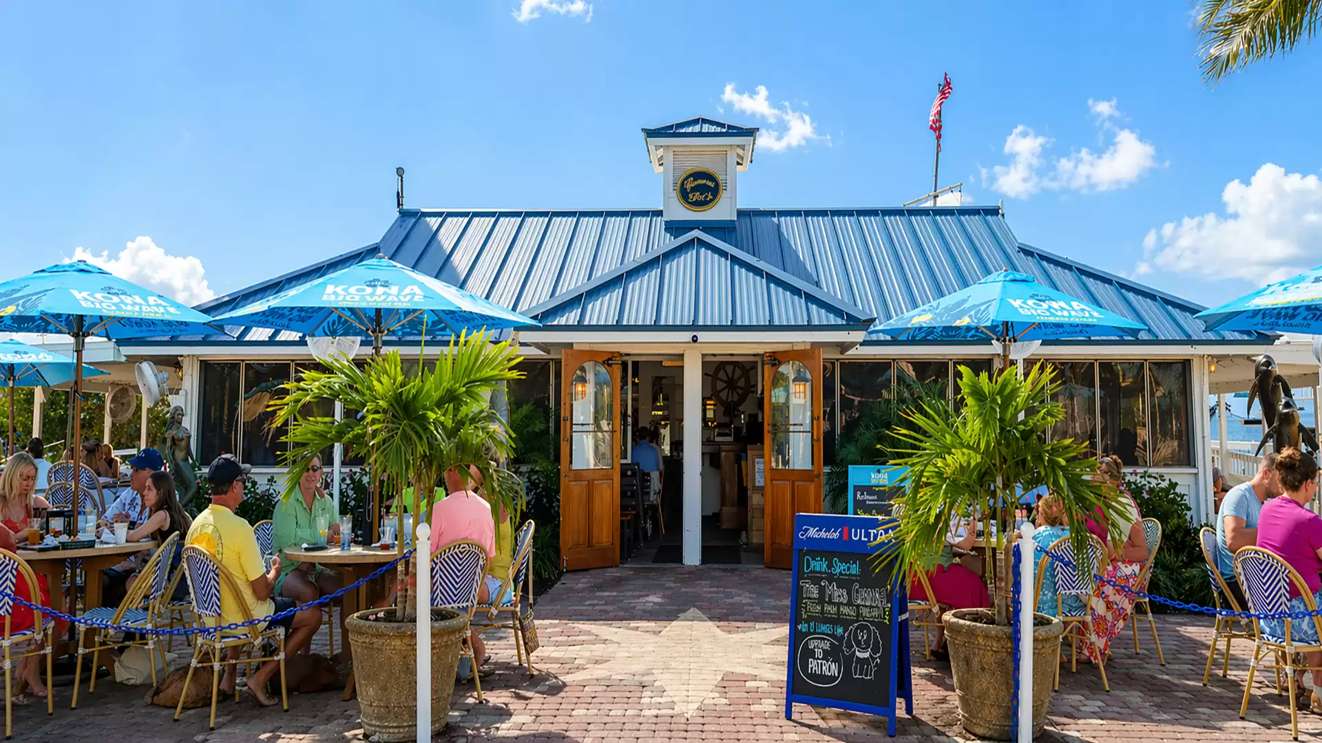 Exterior view of a beach cafe with blue umbrellas, surrounded by seated guests enjoying the sunny weather.