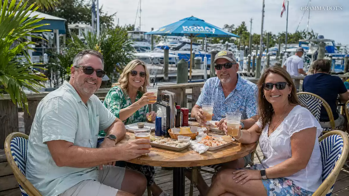 A group of four people sitting at a round outdoor table, enjoying drinks and snacks in a maritime setting with yachts in the background.