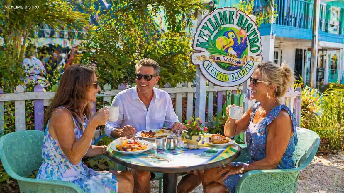 Three people happily enjoying lunch at an outdoor table in a tropical setting with the 'Key Lime Bistro' sign in the background.