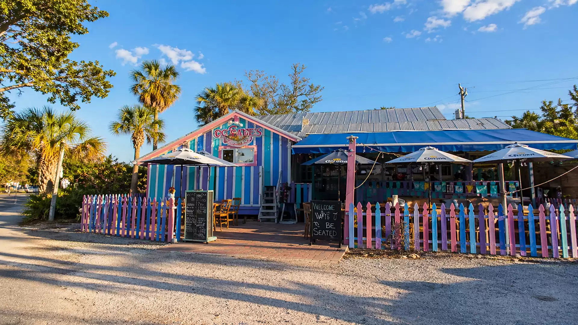 A colorful café with blue and pink striped walls and a colorful fence on a sunny day, surrounded by palm trees.