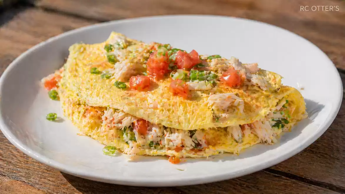 A delicious omelette filled with fresh herbs, tomatoes, and seafood, served on a white plate on a wooden table.