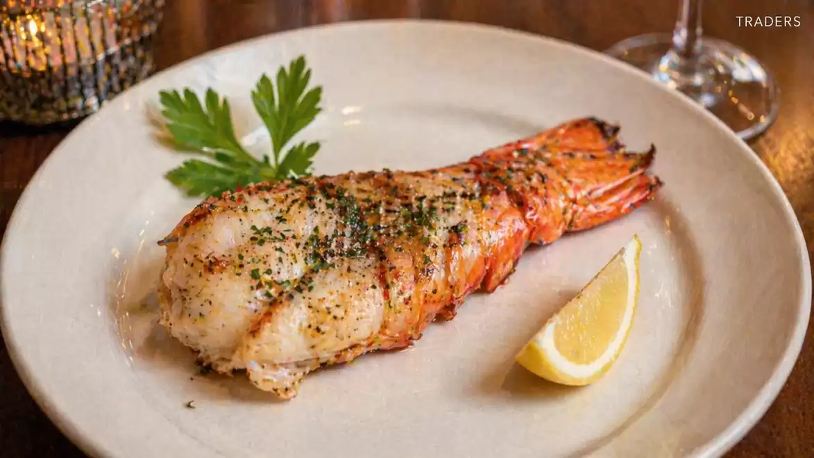 A grilled lobster tail on a white plate, garnished with a lemon wedge and fresh parsley, served on a wooden table.
