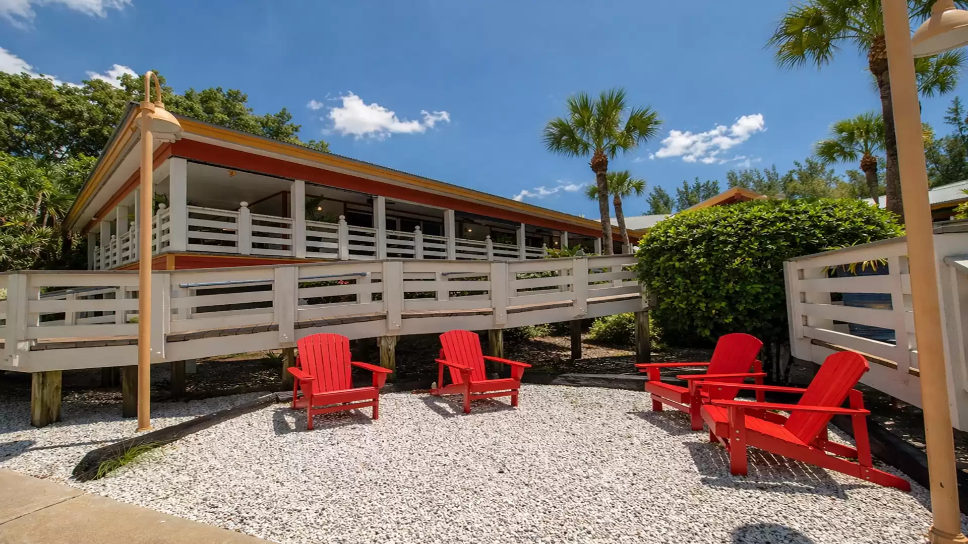 A stilted summer house with white railings, surrounded by green plants and palm trees, with four red garden chairs on gravel under a clear blue sky.