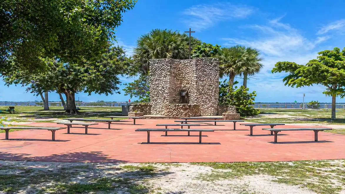 A stone monument with a cross on top, surrounded by palm trees and benches, overlooking the sea in the background.