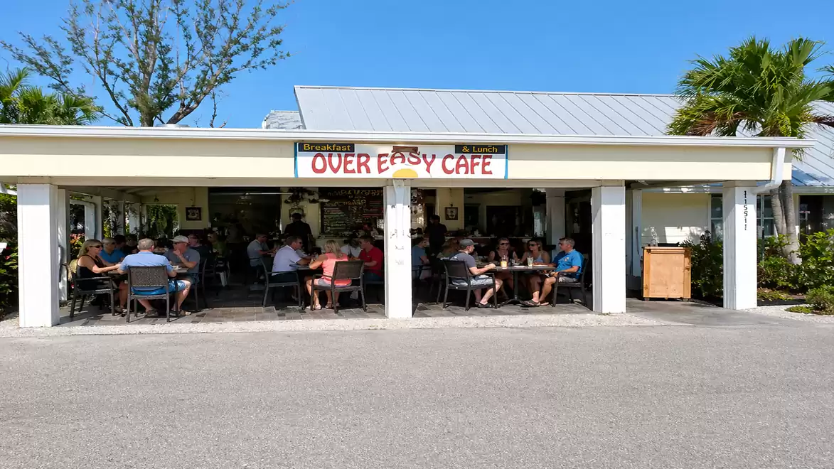 A café called 'Over Easy Cafe' on a sunny day, surrounded by people dining outside, with palm trees in the background.