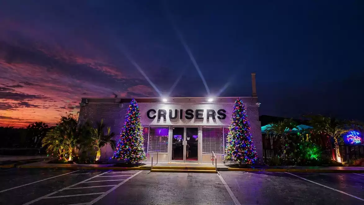The image shows a lit-up building with the sign 'Cruisers'. Two decorated Christmas trees are in front of the entrance. The evening sky in the background displays red and purple colors.